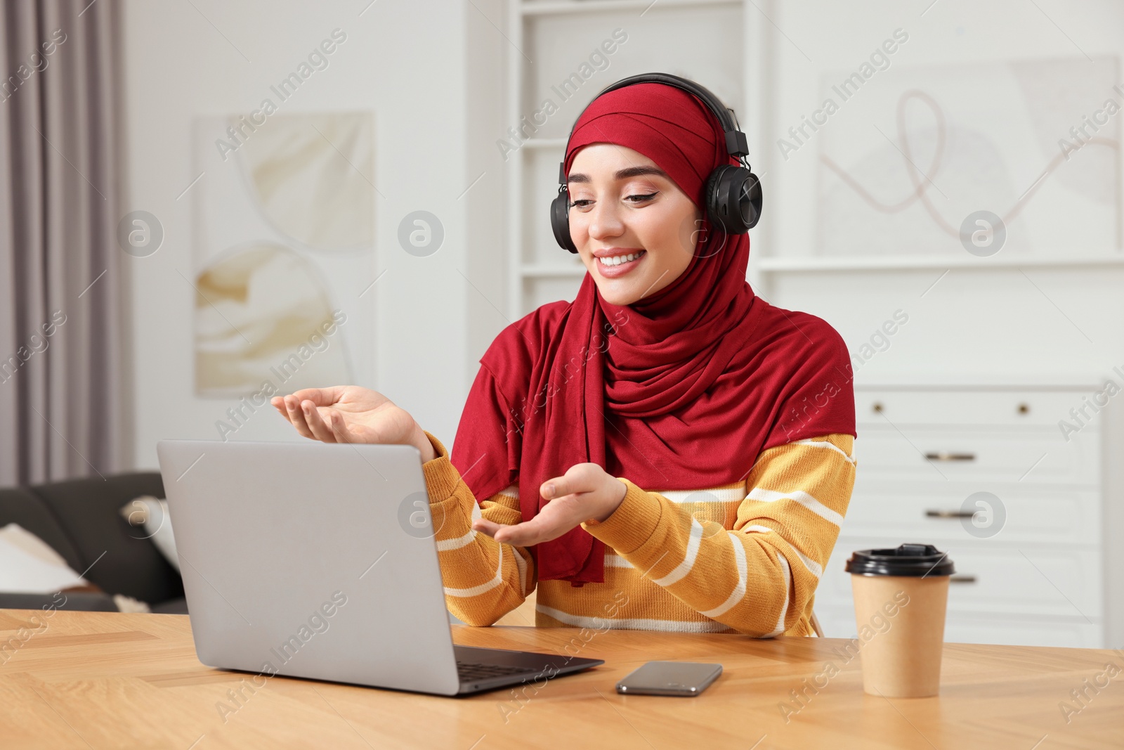 Photo of Muslim woman in hijab using video chat on laptop at wooden table in room