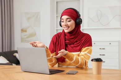 Muslim woman in hijab using video chat on laptop at wooden table in room