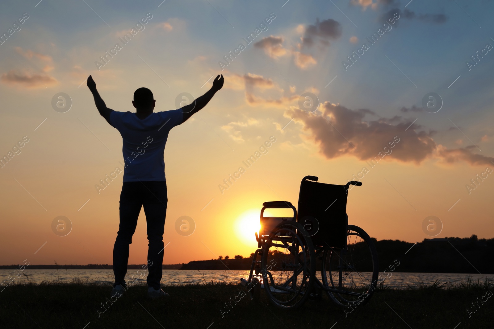 Photo of Man raising hands up to sky near wheelchair at sunset, back view. Healing miracle