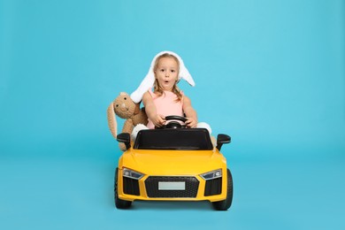 Cute little girl with toy bunny driving children's car on light blue background
