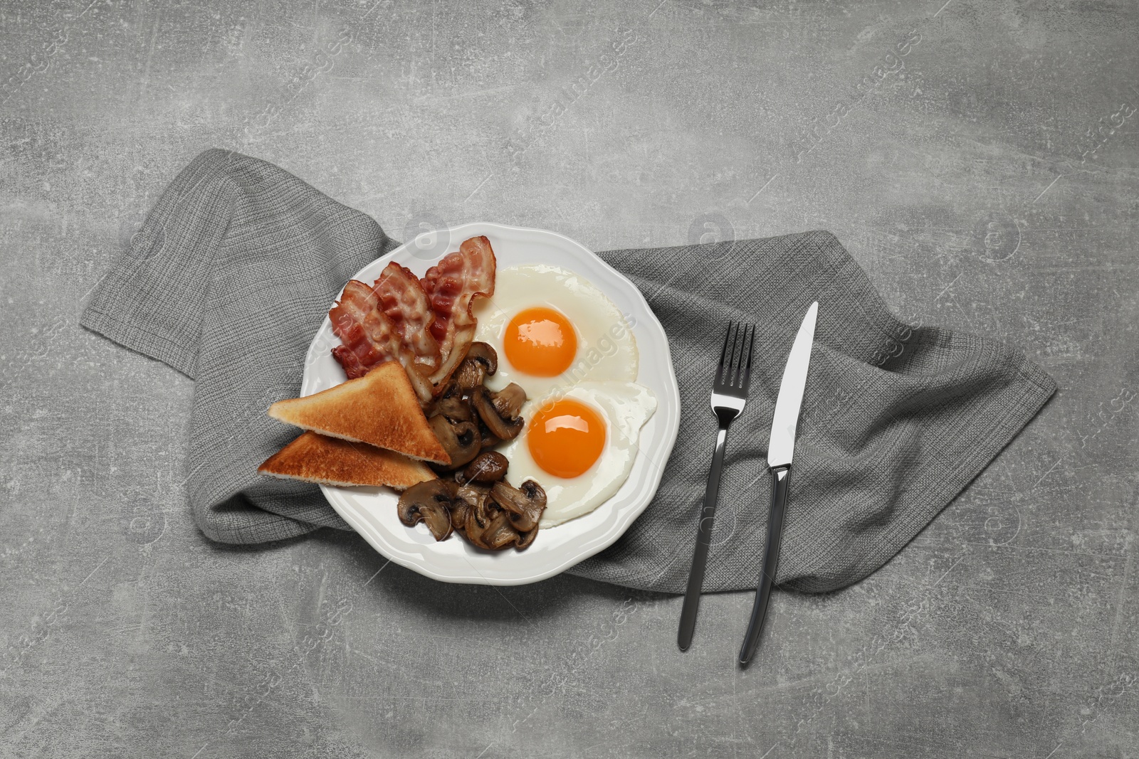 Photo of Plate with fried eggs, mushrooms, bacon and toasted bread on grey table, flat lay. Traditional English breakfast
