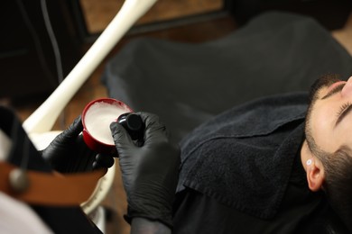 Photo of Professional hairdresser with shaving foam near bearded client in barbershop, closeup