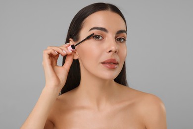 Beautiful young woman applying mascara on grey background