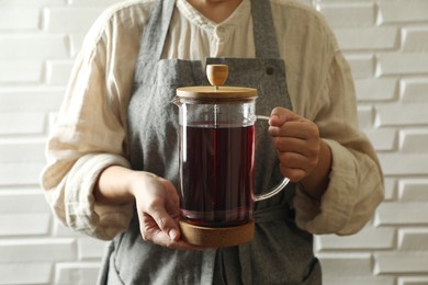 Photo of Woman holding teapot with freshly brewed hibiscus tea near white brick wall, closeup