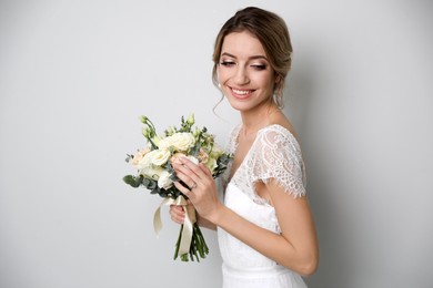 Young bride with elegant hairstyle holding wedding bouquet on light grey background