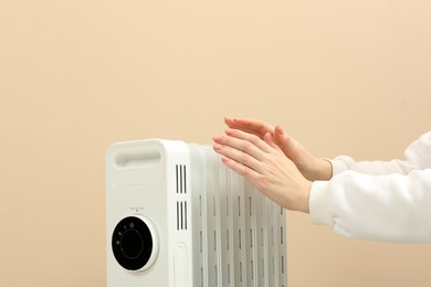 Photo of Young woman warming hands near modern electric heater on beige background, closeup. Space for text