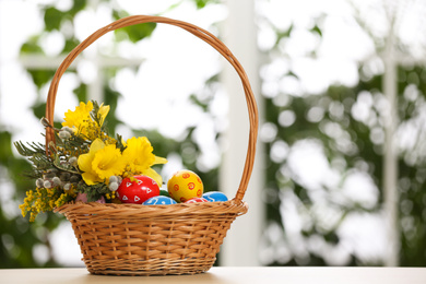 Photo of Easter eggs and bouquet in wicker basket on table against blurred window, space for text