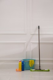 Photo of Floor mop, cleaning detergents, brush and bucket with gloves near white wall indoors