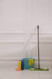 Floor mop, cleaning detergents, brush and bucket with gloves near white wall indoors