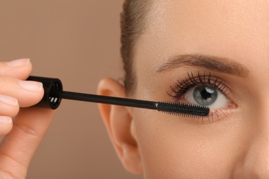 Photo of Woman applying mascara onto eyelashes against light brown background, closeup