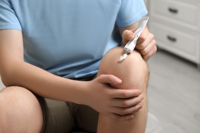Photo of Man applying ointment from tube onto his knee indoors, closeup