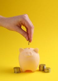 Photo of Financial savings. Woman putting coin into piggy bank on yellow background, closeup