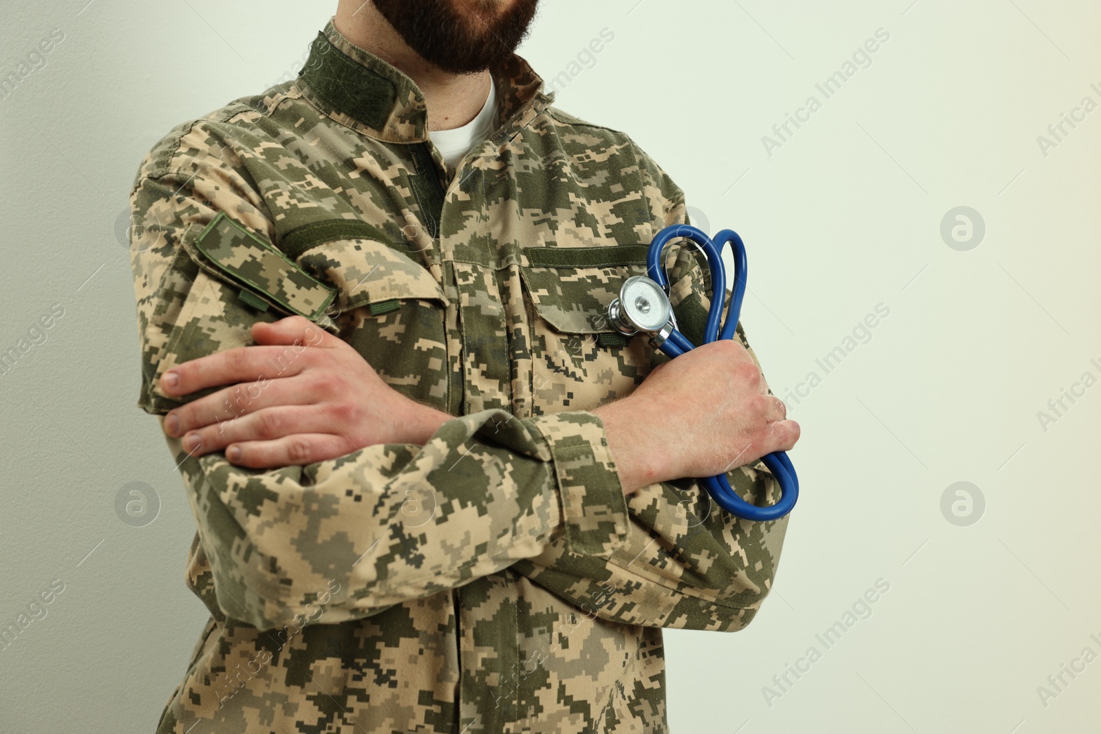 Photo of Man in military uniform with crossed arms and stethoscope on light background, closeup. Health care concept