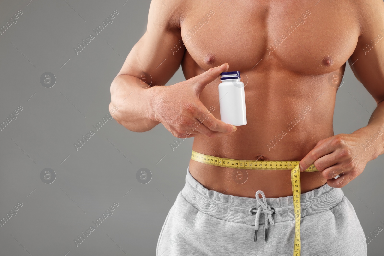 Photo of Weight loss. Athletic man holding bottle of supplements and measuring his waist with tape on grey background, closeup. Space for text