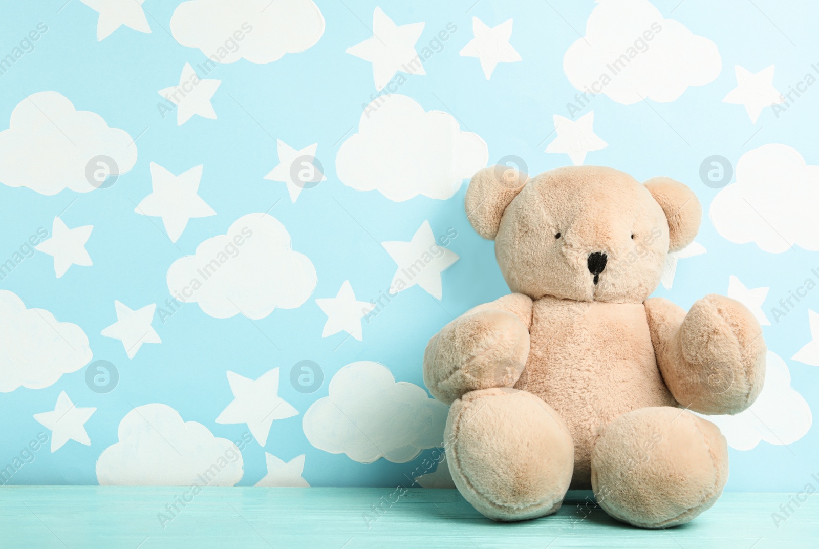 Photo of Teddy bear on wooden table near wall with painted blue sky, space for text. Baby room interior