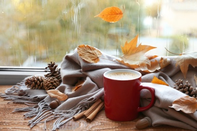 Photo of Cup of hot drink, scarf and autumn leaves on windowsill. Cozy atmosphere