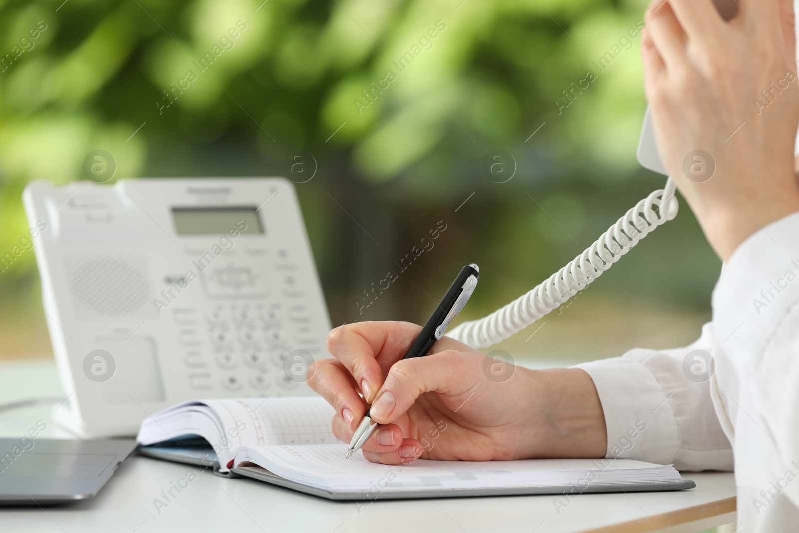 Photo of Assistant with telephone handset writing at white table against blurred green background, closeup