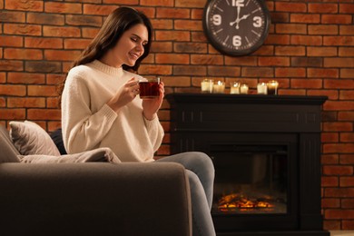 Photo of Young woman with cup of tea relaxing in armchair near fireplace at home