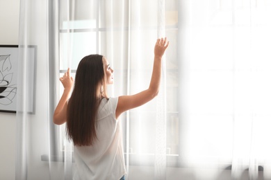 Young woman opening window curtains at home. Space for text