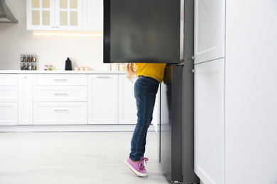 Photo of Little girl looking into open refrigerator in kitchen. Space for text