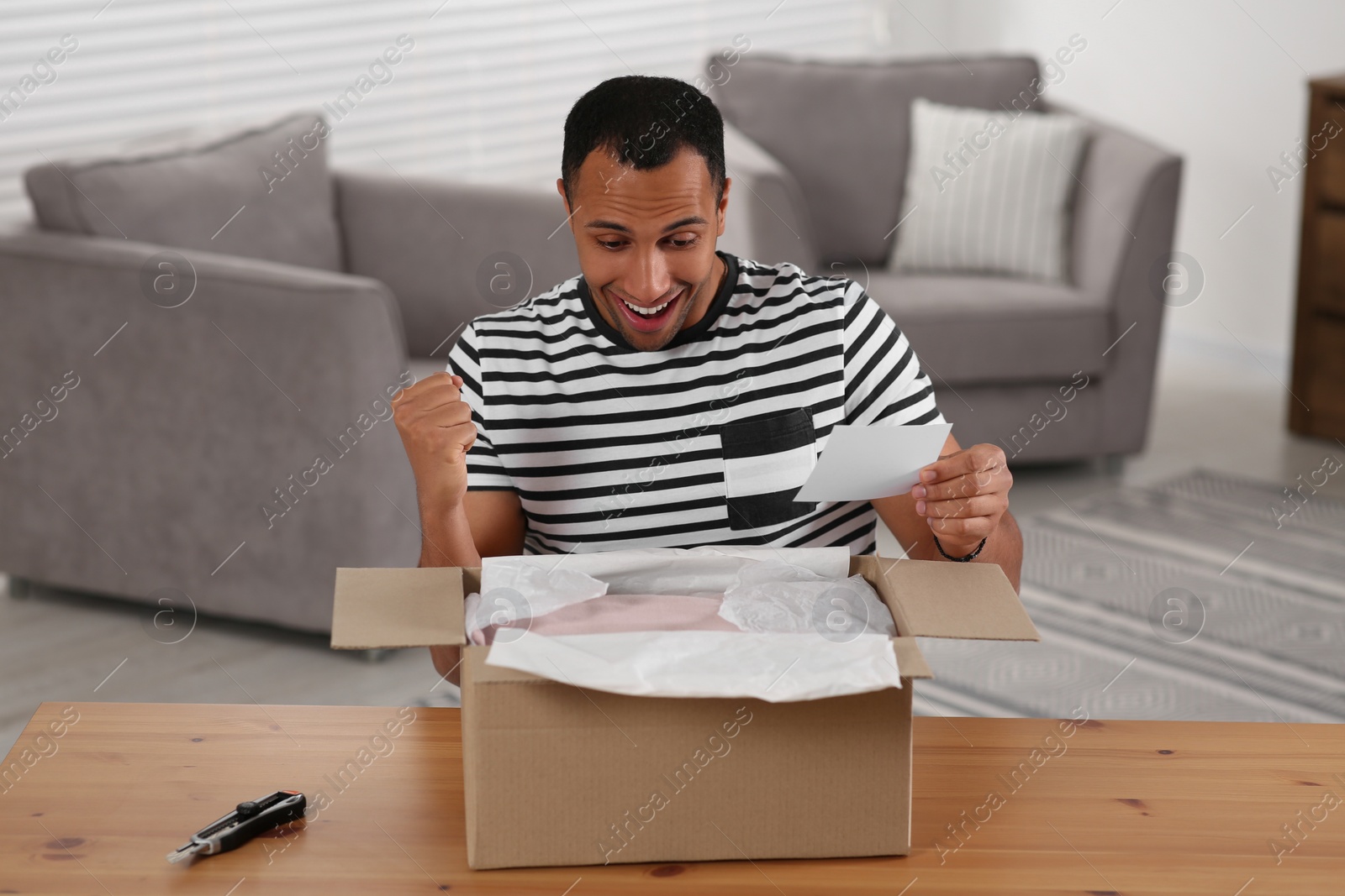 Photo of Emotional young man with greeting card near parcel at table indoors. Internet shopping