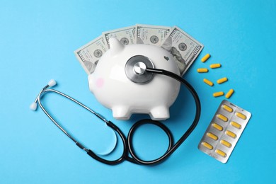Photo of White ceramic piggy bank, stethoscope, money and pills on light blue background, flat lay. Medical insurance