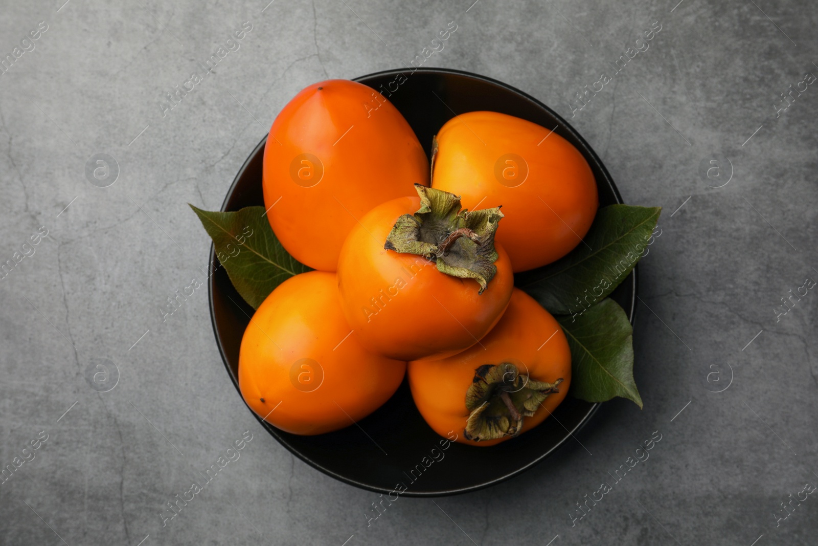Photo of Delicious ripe persimmons in bowl on light gray textured table, top view