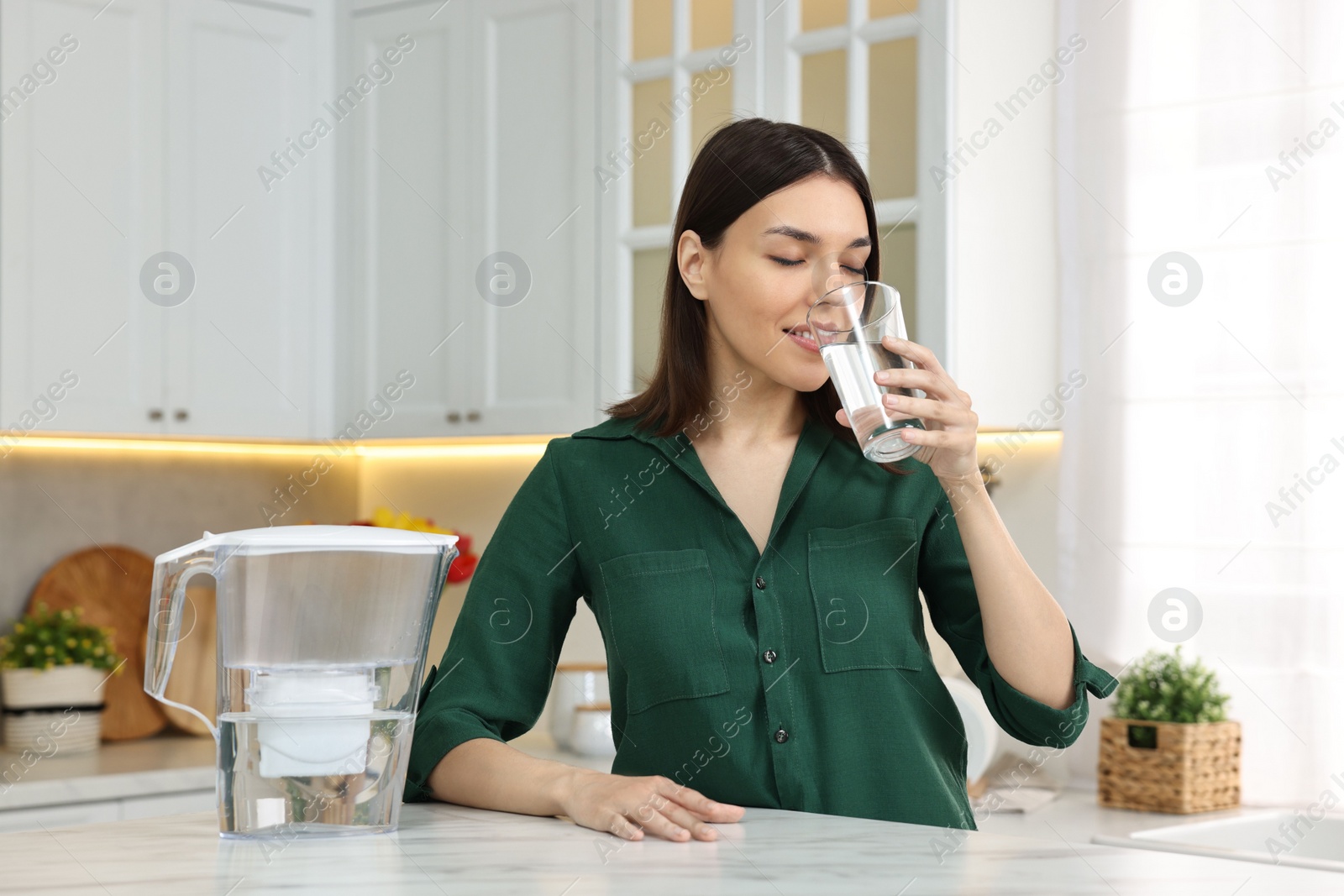 Photo of Woman drinking water and filter jug in kitchen