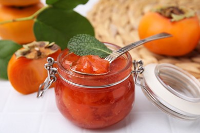 Jar and spoon of tasty persimmon jam, ingredients on white tiled table, closeup