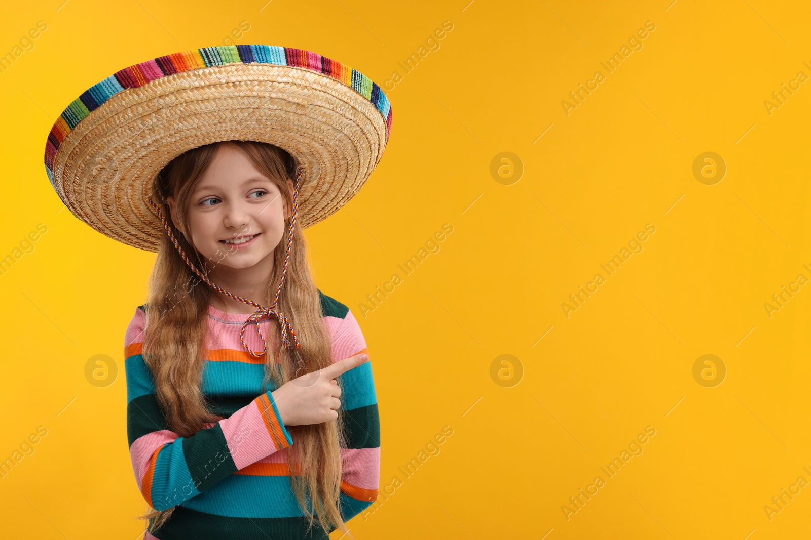 Photo of Cute girl in Mexican sombrero hat pointing at something on orange background. Space for text