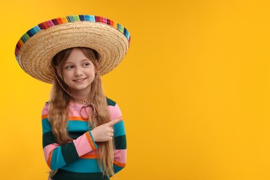 Photo of Cute girl in Mexican sombrero hat pointing at something on orange background. Space for text