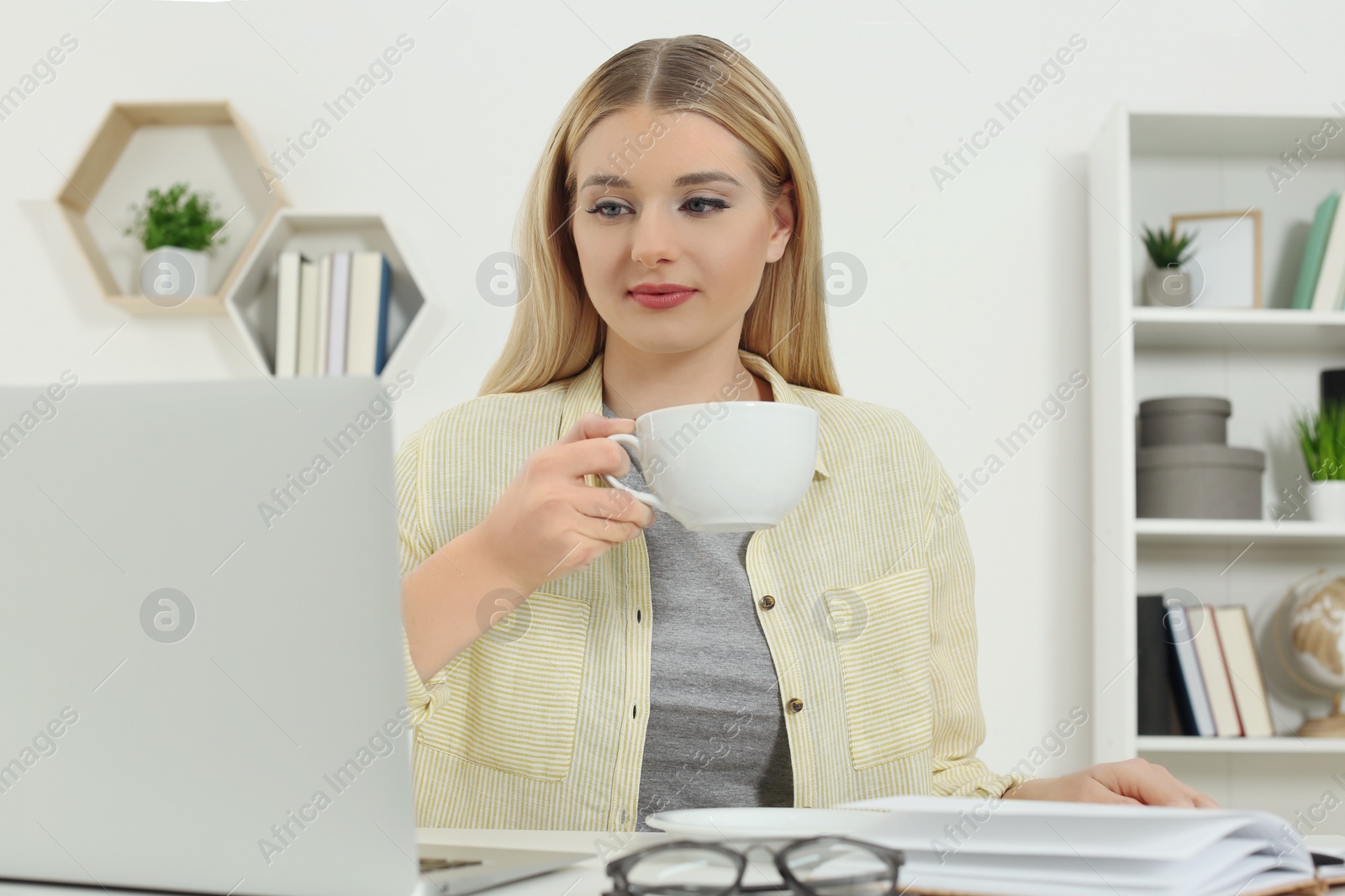 Photo of Home workplace. Woman with cup of hot drink working on laptop at white desk in room