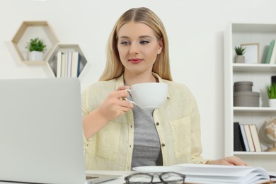 Photo of Home workplace. Woman with cup of hot drink working on laptop at white desk in room