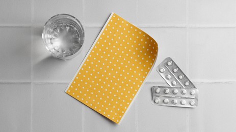 Photo of Pepper plaster, pills and glass of water on white tiled table, flat lay