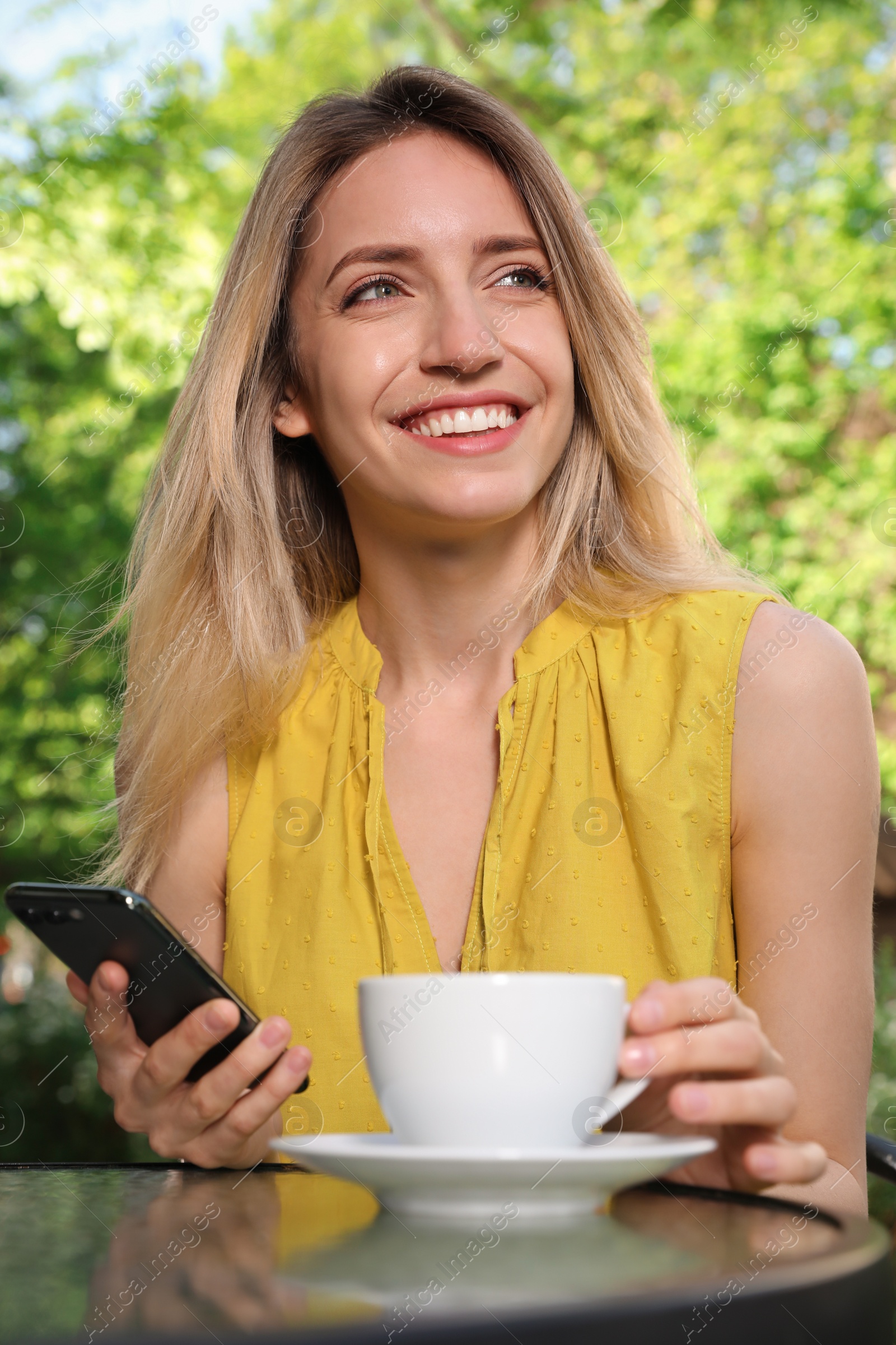 Photo of Happy young woman with cup of coffee and smartphone enjoying early morning in outdoor cafe