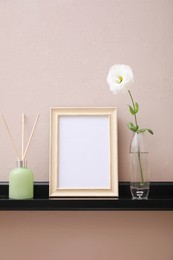 Photo of Empty photo frame, air reed freshener and beautiful flower on shelf near pink wall