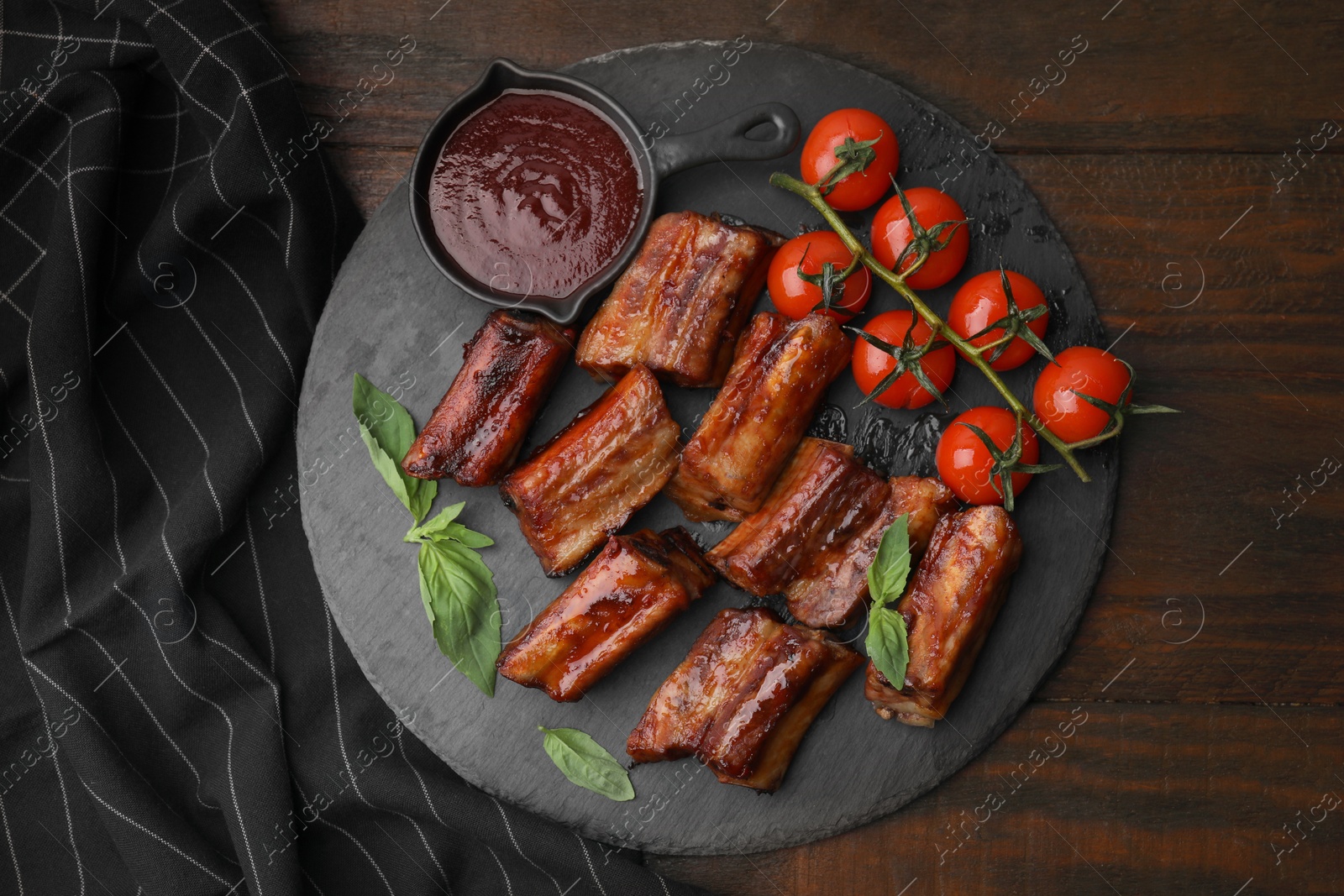 Photo of Tasty roasted pork ribs served with sauce, basil and tomatoes on wooden table, top view