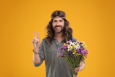 Photo of Hippie man with bouquet of colorful flowers showing V-sign on orange background