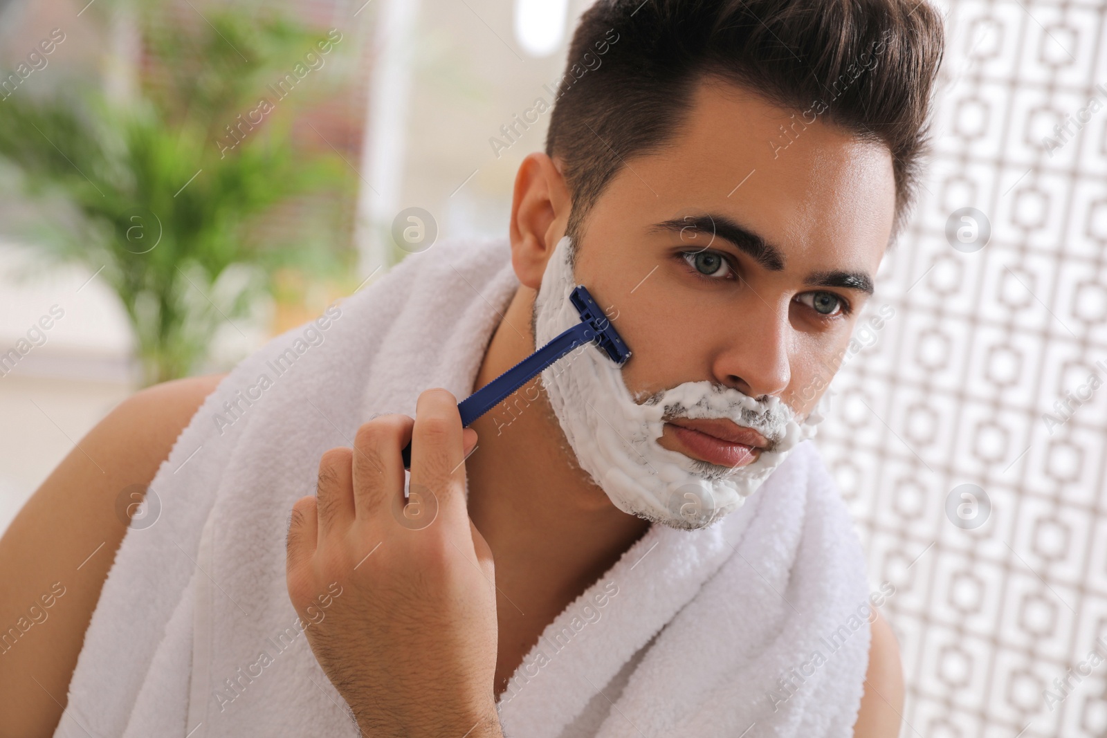 Photo of Handsome young man shaving with razor in bathroom