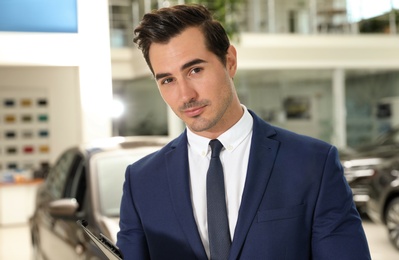 Young salesman with clipboard near car in modern dealership