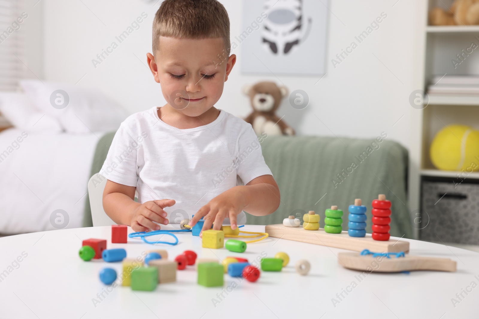 Photo of Motor skills development. Little boy playing with wooden pieces and string for threading activity at white table indoors