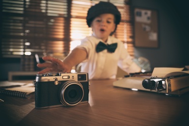 Cute little detective taking camera at table in office, focus on device