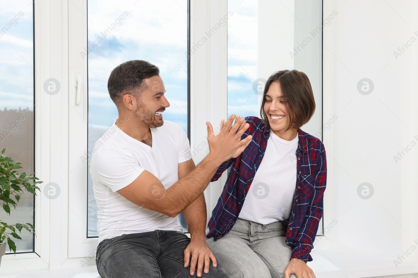 Photo of Happy couple giving high five while sitting on windowsill in new apartment. Moving day