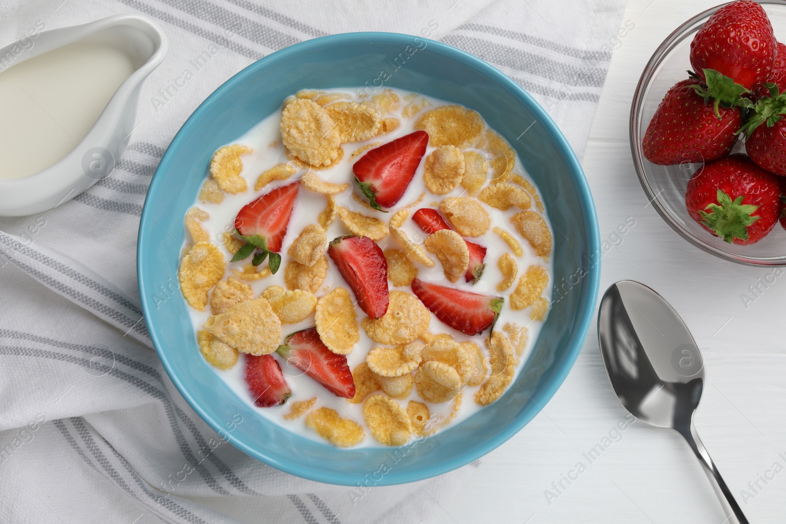 Photo of Bowl of tasty crispy corn flakes with milk and strawberries on white wooden table, flat lay