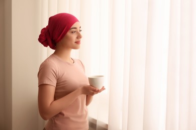 Photo of Cancer patient. Young woman with headscarf and hot drink near window indoors, space for text