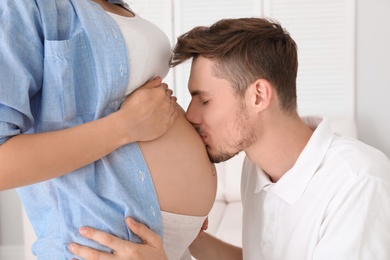 Photo of Young husband kissing his pregnant wife's tummy at home