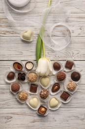 Heart made with delicious chocolate candies and beautiful tulip on white wooden table, flat lay