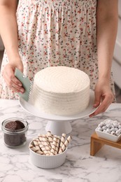 Photo of Woman using scraper to decorate cake at white marble table, closeup