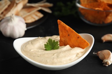 Photo of Delicious hummus with nacho chip and parsley on black table, closeup