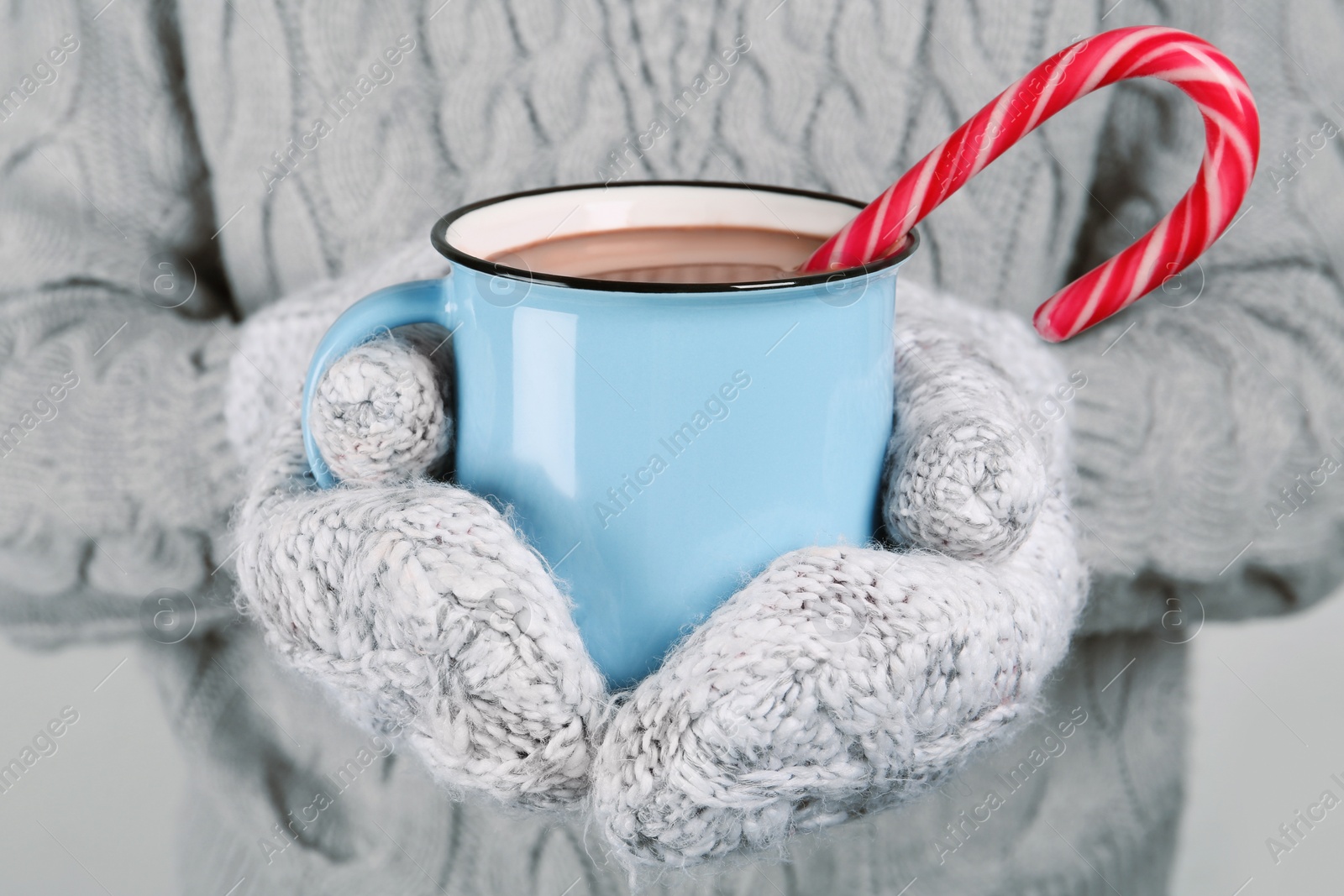 Photo of Woman in knitted mittens holding cup of delicious hot chocolate with candy cane, closeup